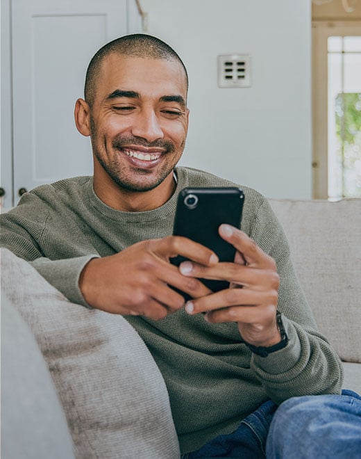 A young man sitting casually on a couch smiles while holding his smart phone.