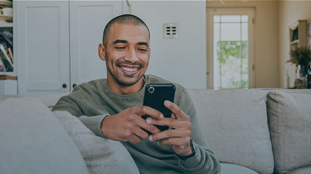 A young man sitting casually on a couch smiles while holding his smart phone.